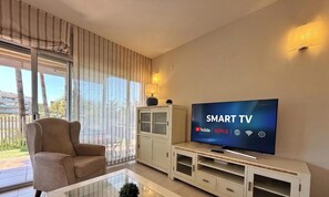 A modern living room with a large flat-screen TV displaying 'SMART TV' and streaming service logos, a beige sofa, a glass coffee table, and a sliding glass door leading to a balcony.