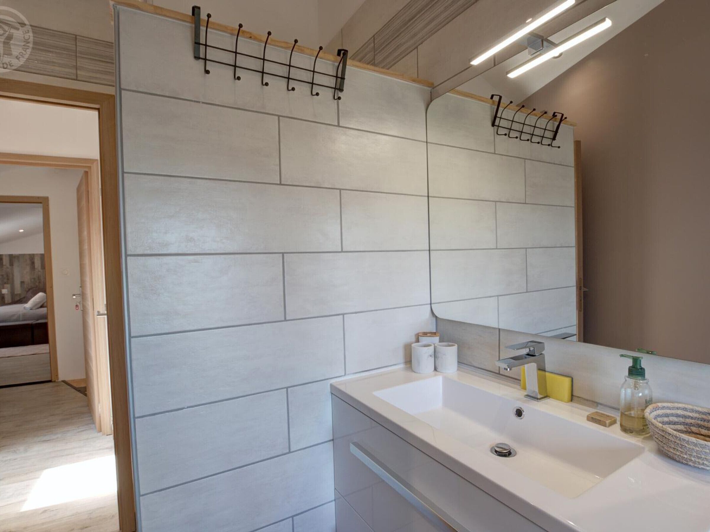 A modern bathroom with a large mirror, white tiles, a sink with toiletries, and a wooden door.
