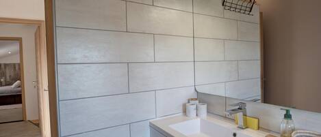 A modern bathroom with a large mirror, white tiles, a sink with toiletries, and a wooden door.