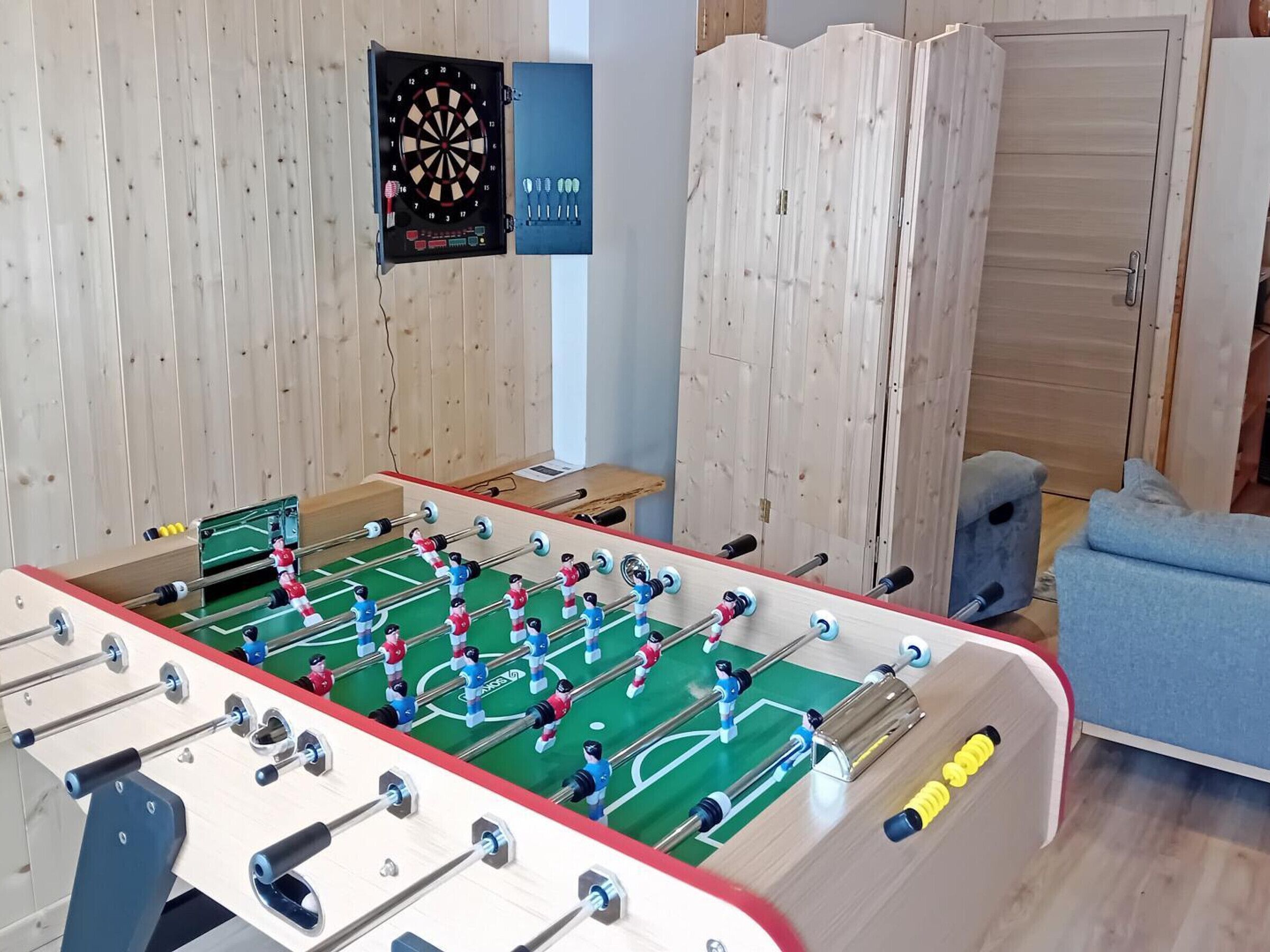 Ein Fußballtisch in einem Raum mit Holzwänden, einer Dartscheibe und einem blauen Sofa.