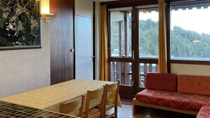 Une pièce comprenant une table à manger, des chaises, un canapé, un tableau et une grande fenêtre donnant sur une zone boisée.