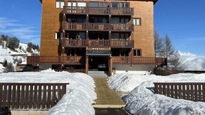 Un bâtiment en bois à plusieurs étages, doté de balcons, avec une enseigne indiquant « MUSTAGO », et un paysage enneigé en arrière-plan.