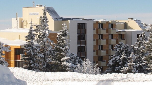 A modern building with a sign reading "Edelweiss." It features a mix of residential and commercial spaces with a flat roof and snow-covered trees in the foreground.