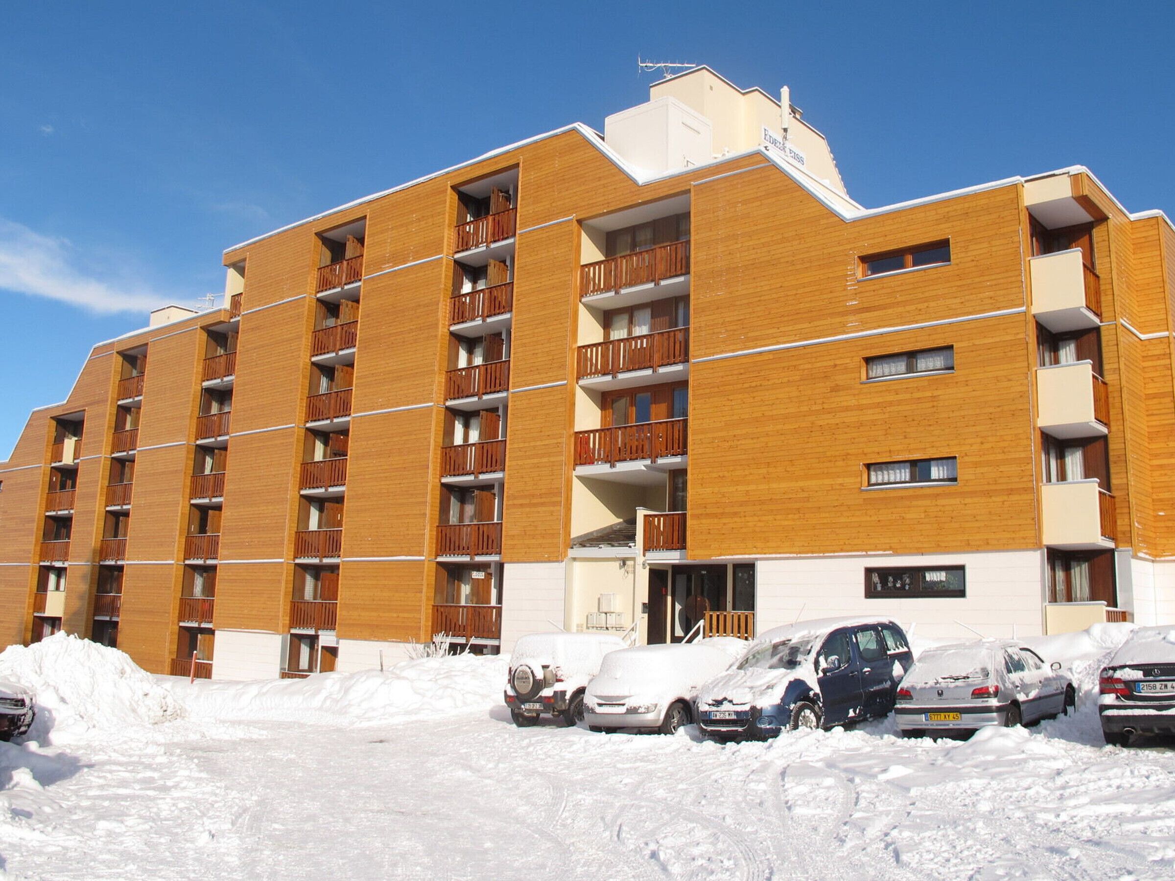 Un hotel de varios pisos con fachada de madera, balcones y un estacionamiento cubierto de nieve.
