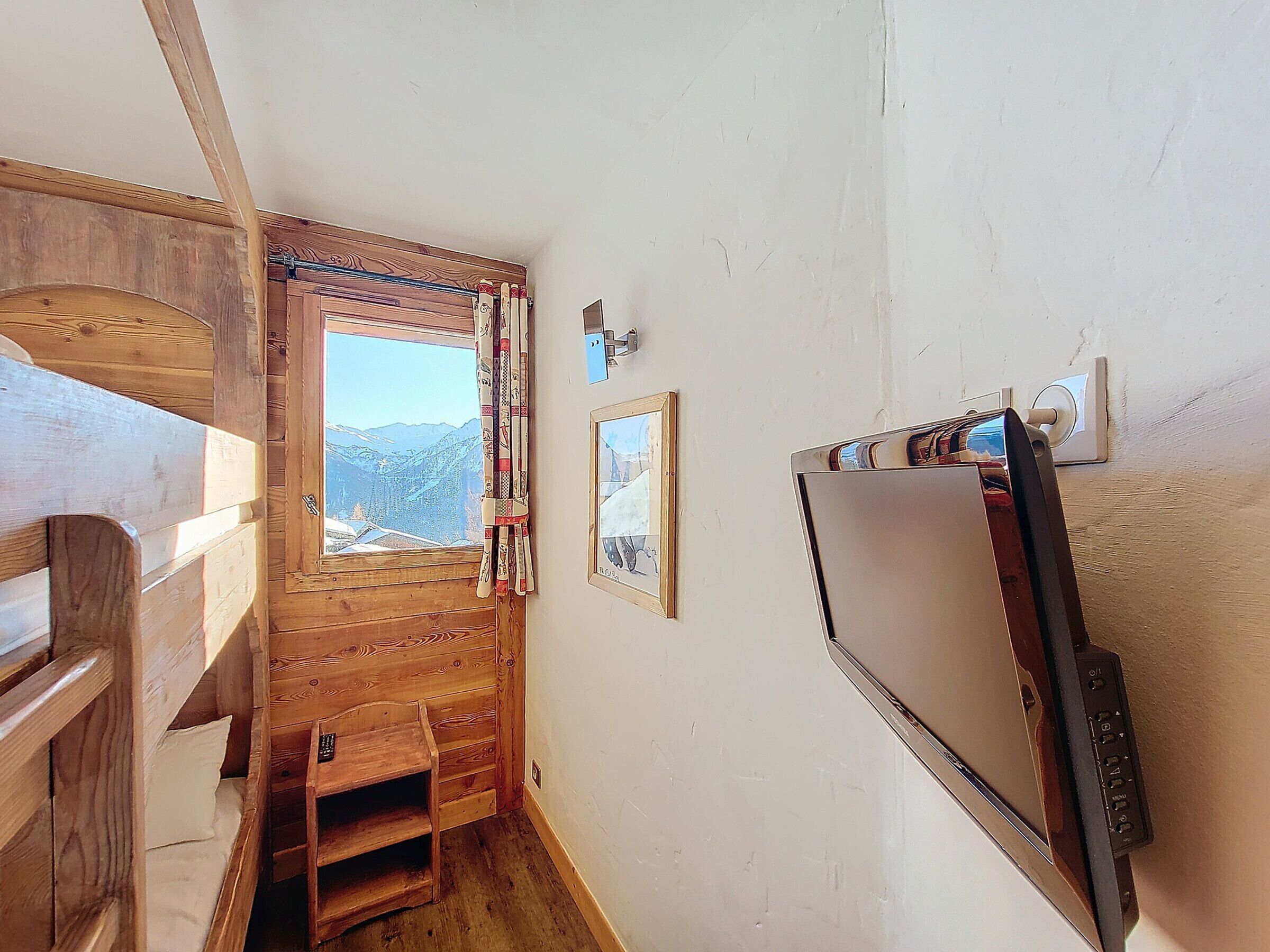 A wooden cabin interior with a window offering a mountain view, a bed, a small desk, and a mounted television.