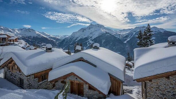 Un villaggio montano innevato con case di legno rustiche e una vista chiara delle cime circostanti.