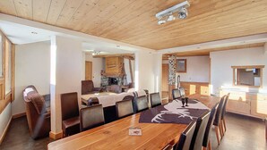 Un'ampia sala da pranzo con un lungo tavolo in legno e numerose sedie, un camino in pietra e una cucina visibile attraverso un'apertura.