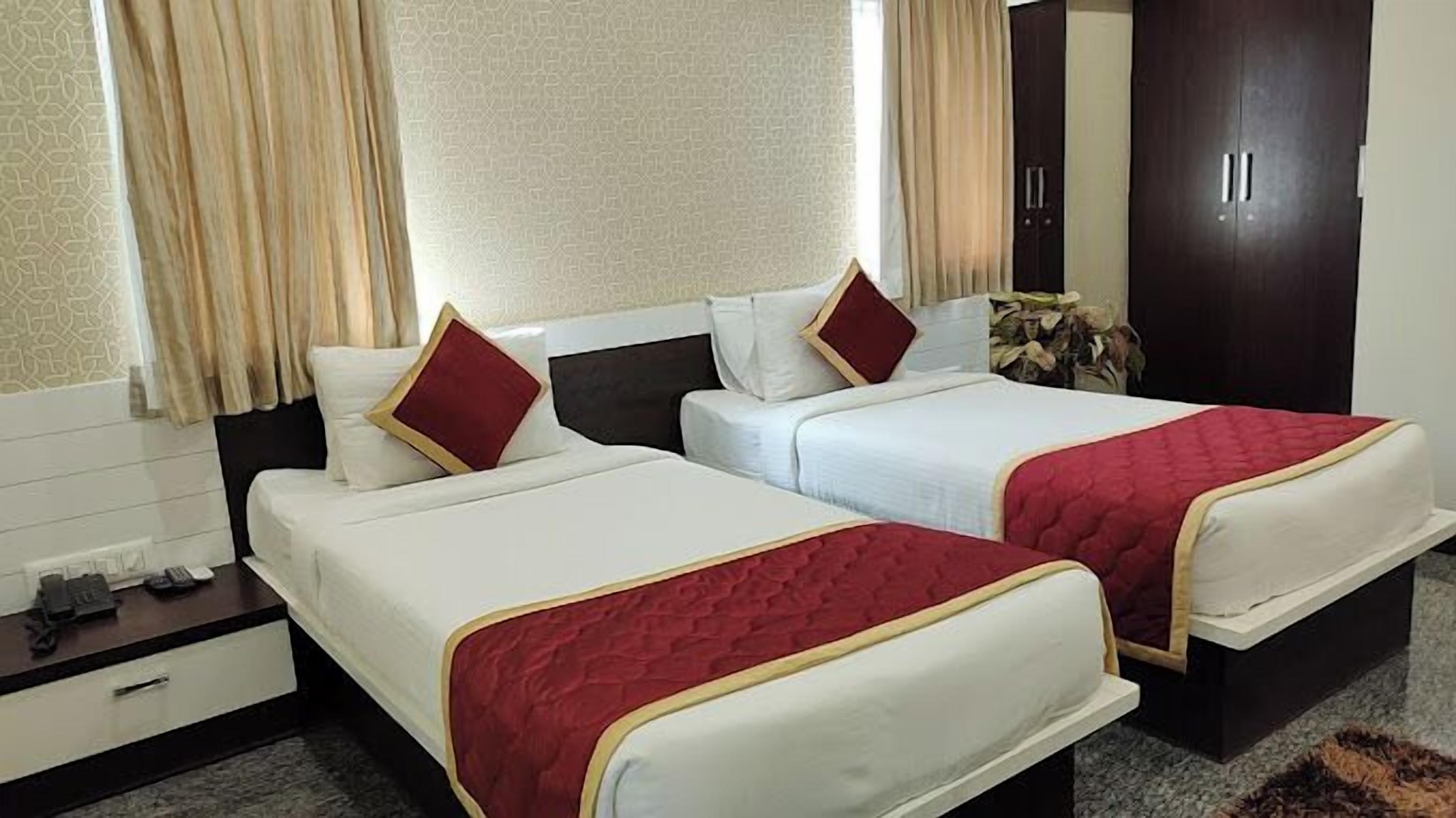 Habitació d'hotel amb dos llits, cadascun amb roba de llit blanca i detalls vermells, un calaixer de fusta, un armari i una finestra amb cortines.