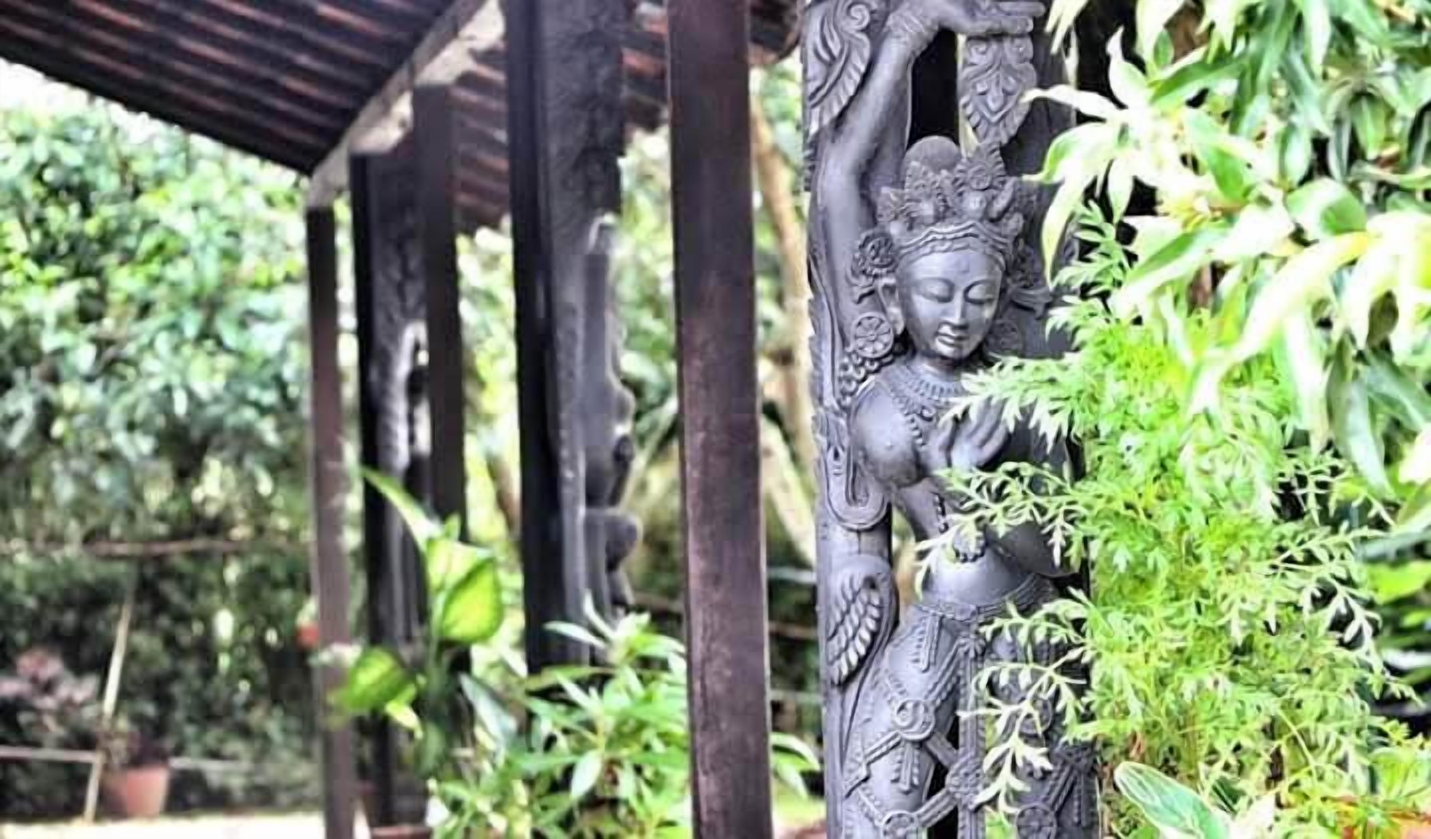 Una estatua detallada con tallas intrincadas, frente a un telón de fondo de vegetación exuberante y una estructura de madera.