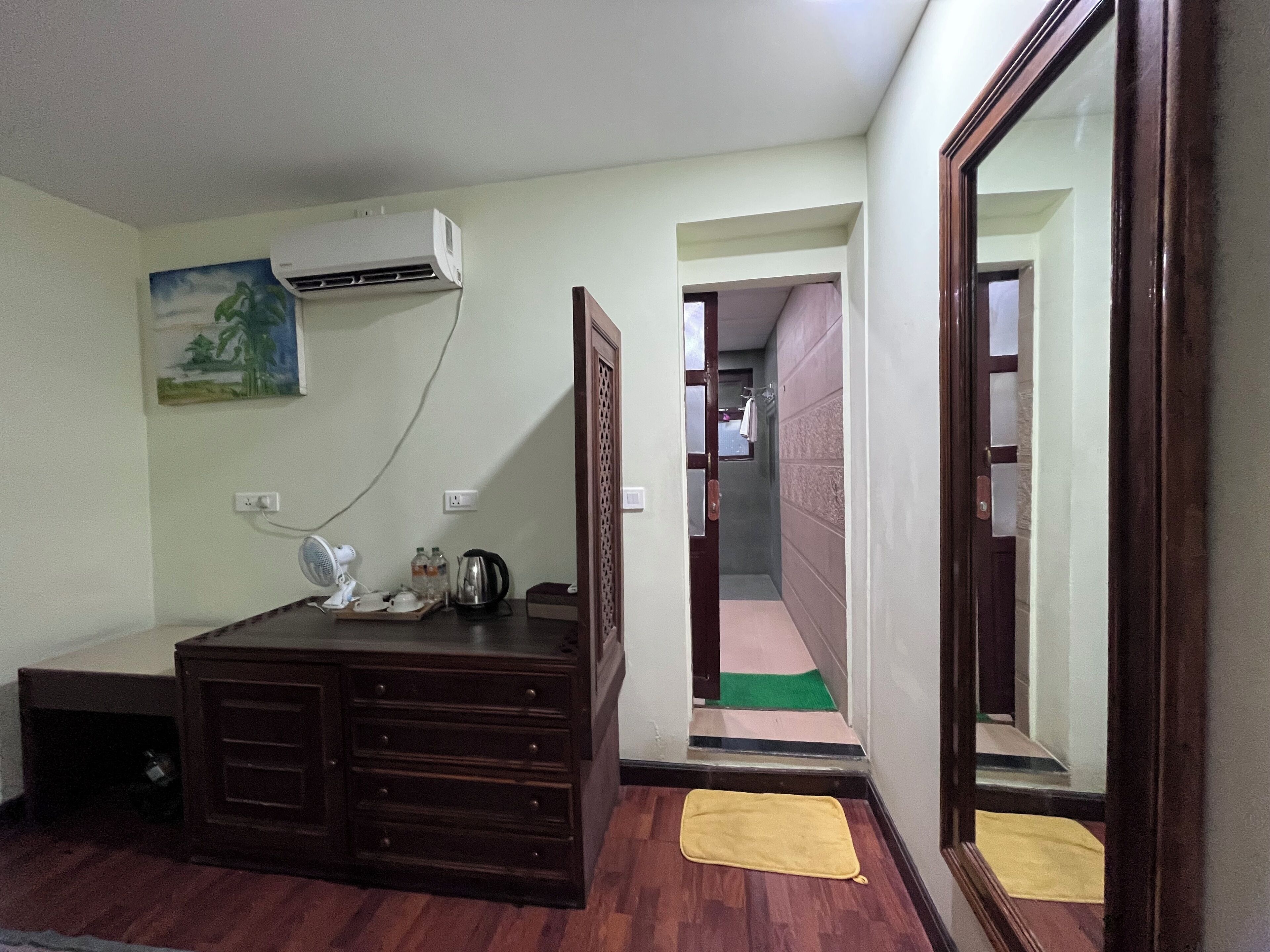 Habitación de hotel con cómoda de madera, equipo de aire acondicionado, ventilador, un cuadro y baño visible a través de una puerta abierta.