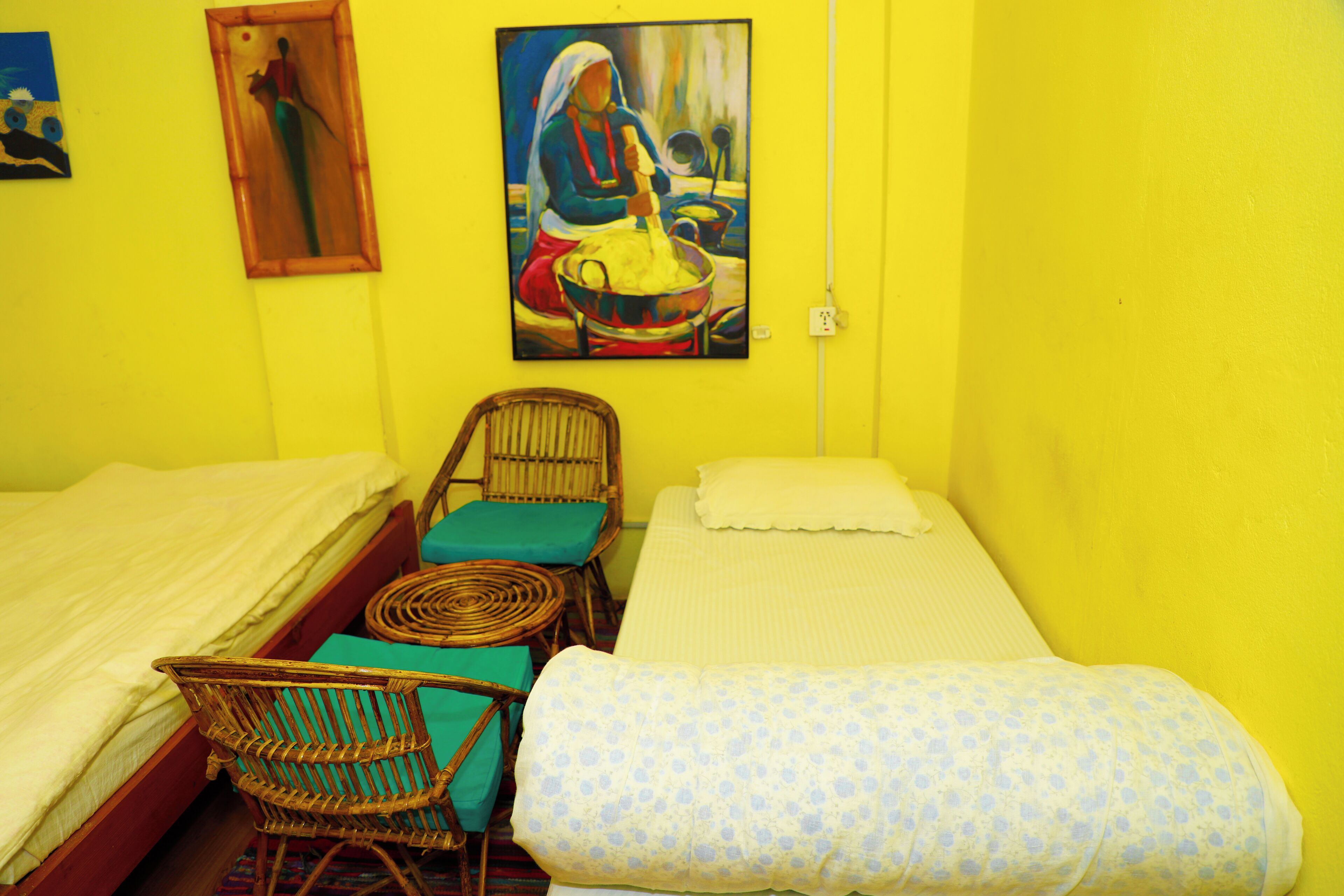 A room with two beds, wicker chairs, a painting, and a yellow wall.
