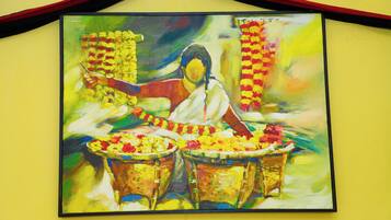 A painting of a woman playing a drum, surrounded by flowers and draped in a white sari, against a yellow wall.