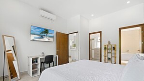Une chambre à coucher avec un lit, un bureau, une chaise, un téléviseur et une salle de bain visible à travers une porte ouverte.