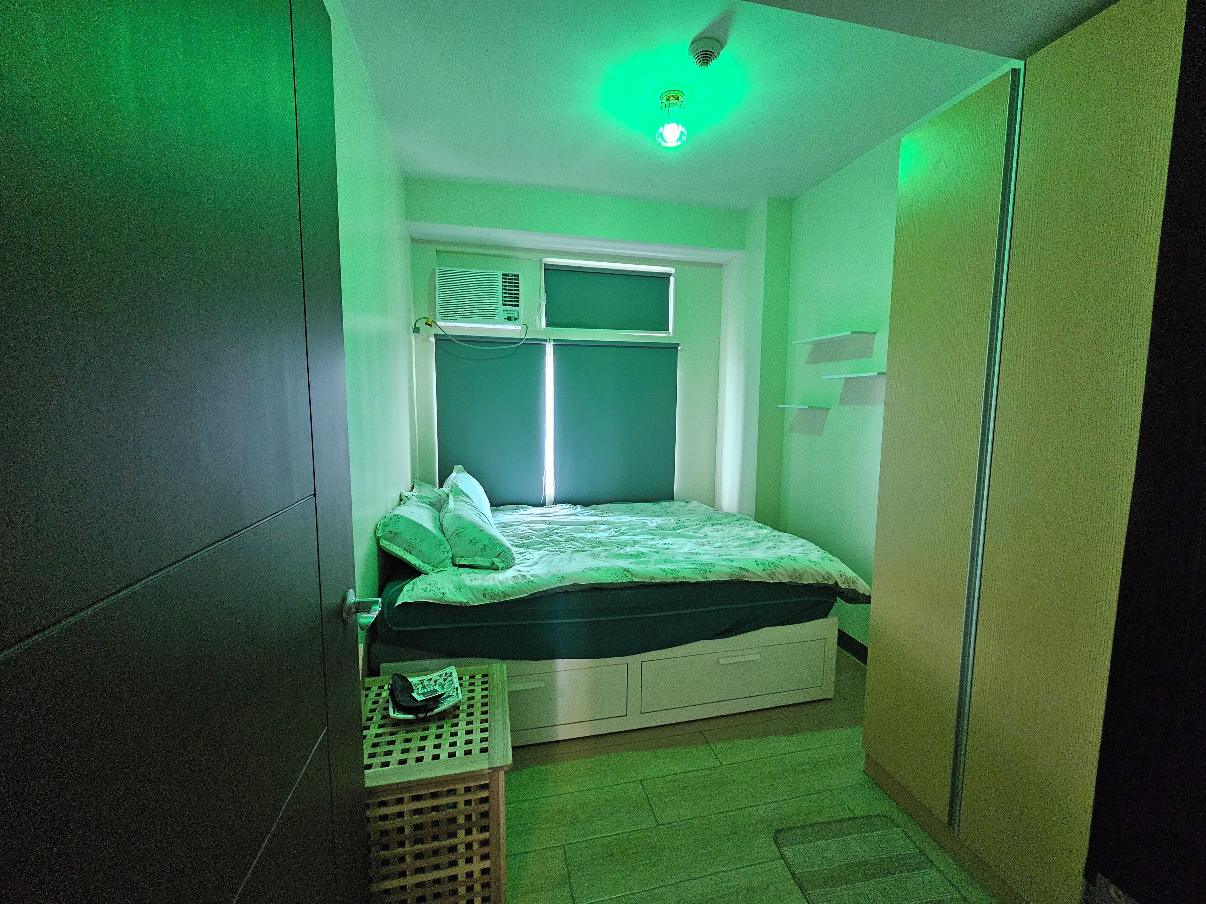 Une petite pièce étroite, avec un lit, une fenêtre et un luminaire au plafond.
