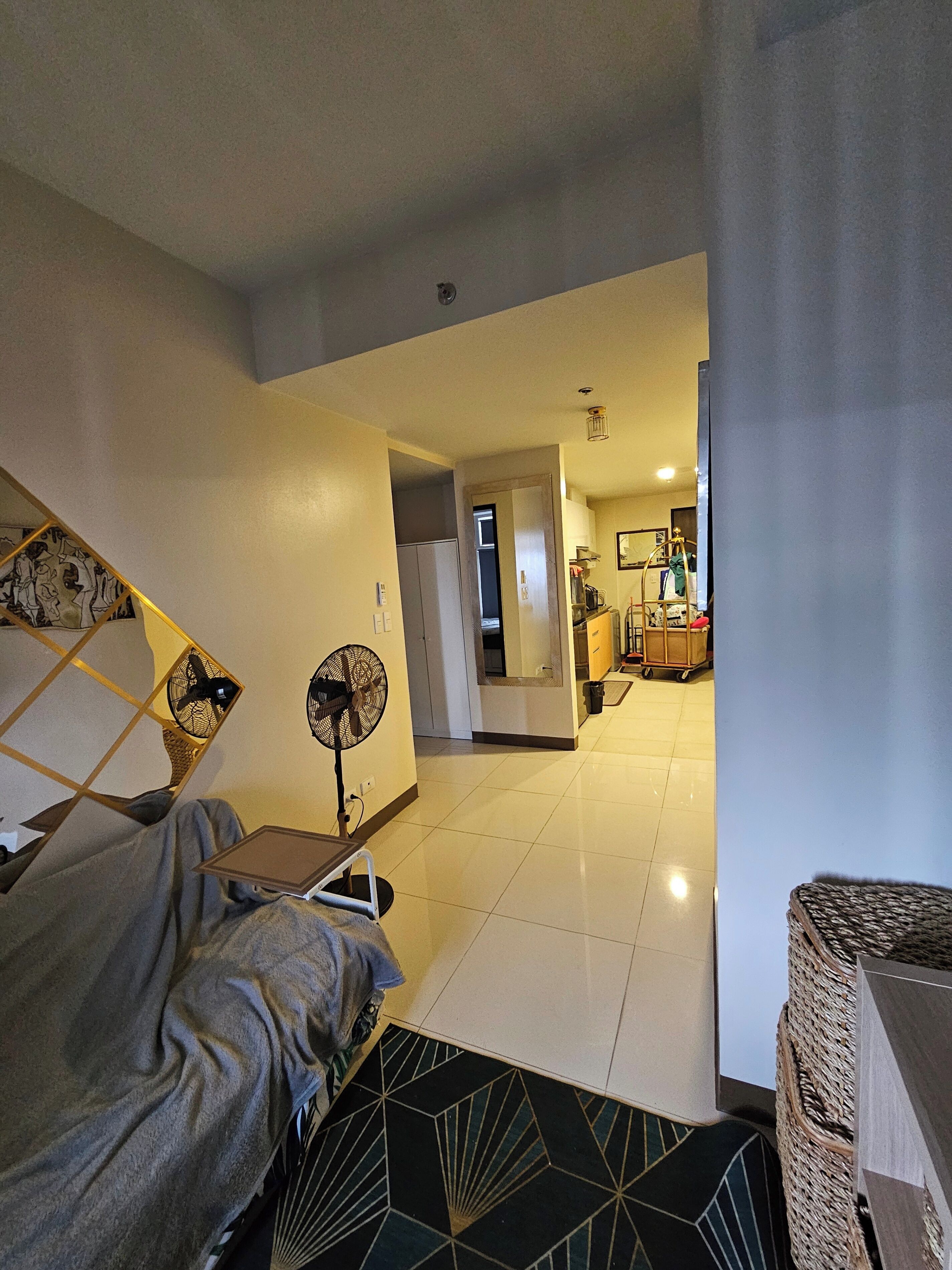 A hotel room with a bed, a standing fan, a small table, and a view into the kitchen area.