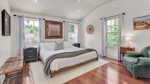 A spacious bedroom with a large bed, a bench, and a green armchair. There are two windows with curtains, a ceiling fan, and a painting on the wall.