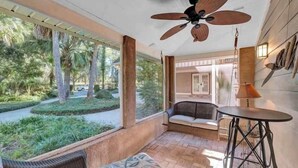 A sunlit patio with a ceiling fan, a cushioned seating area, and a view of a landscaped garden.