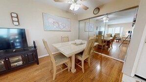 Un espace repas séparé par une cloison en verre, avec des chaises en bois, une table blanche et un téléviseur posé sur un meuble.