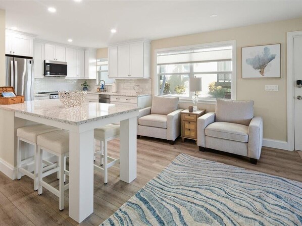 A modern kitchen with white cabinetry, a central island with bar stools, a dining area with a sofa, and a large window providing natural light.