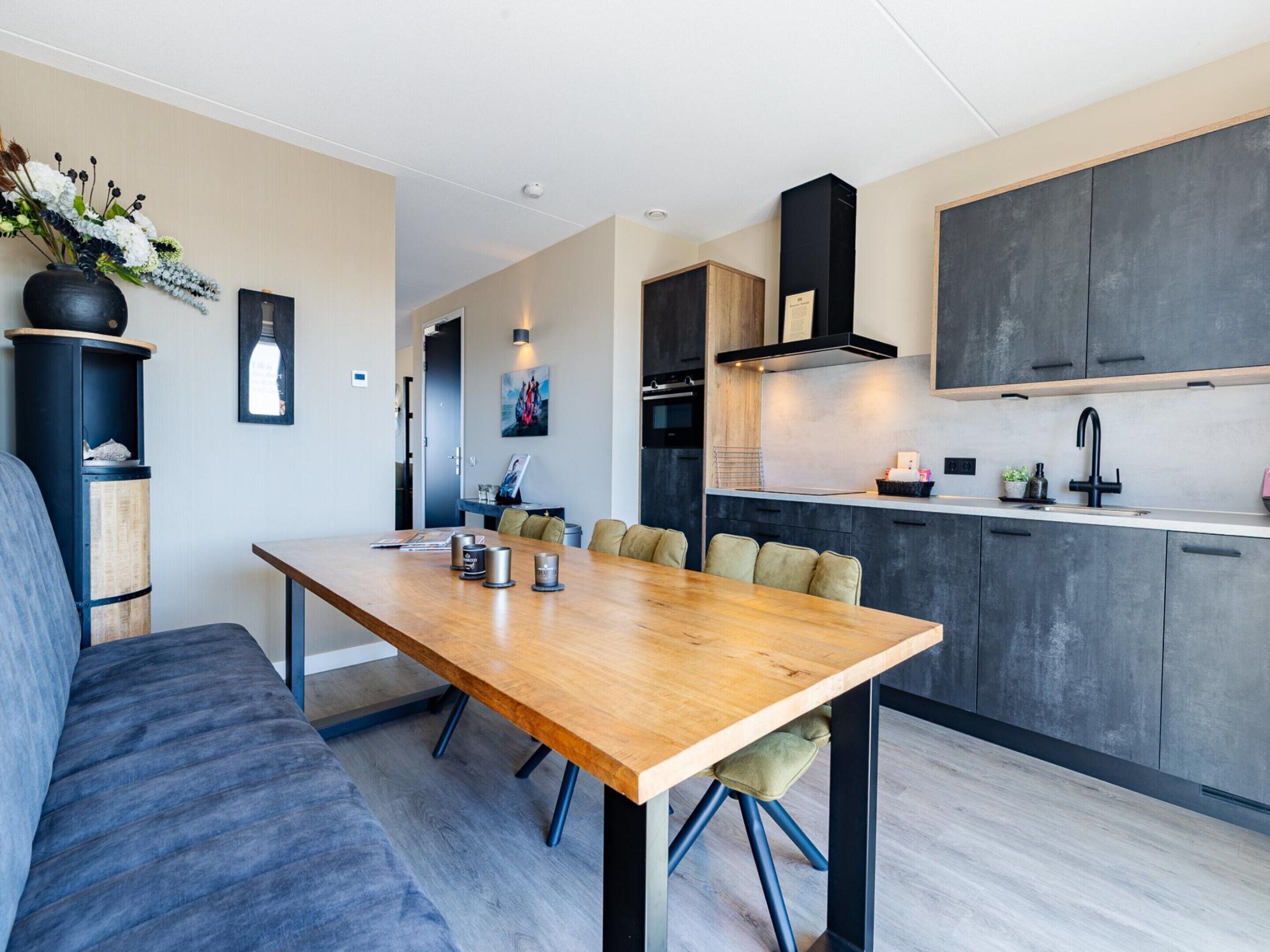 Een moderne keuken met donkere kastjes, een houten eettafel en een grijze bank.