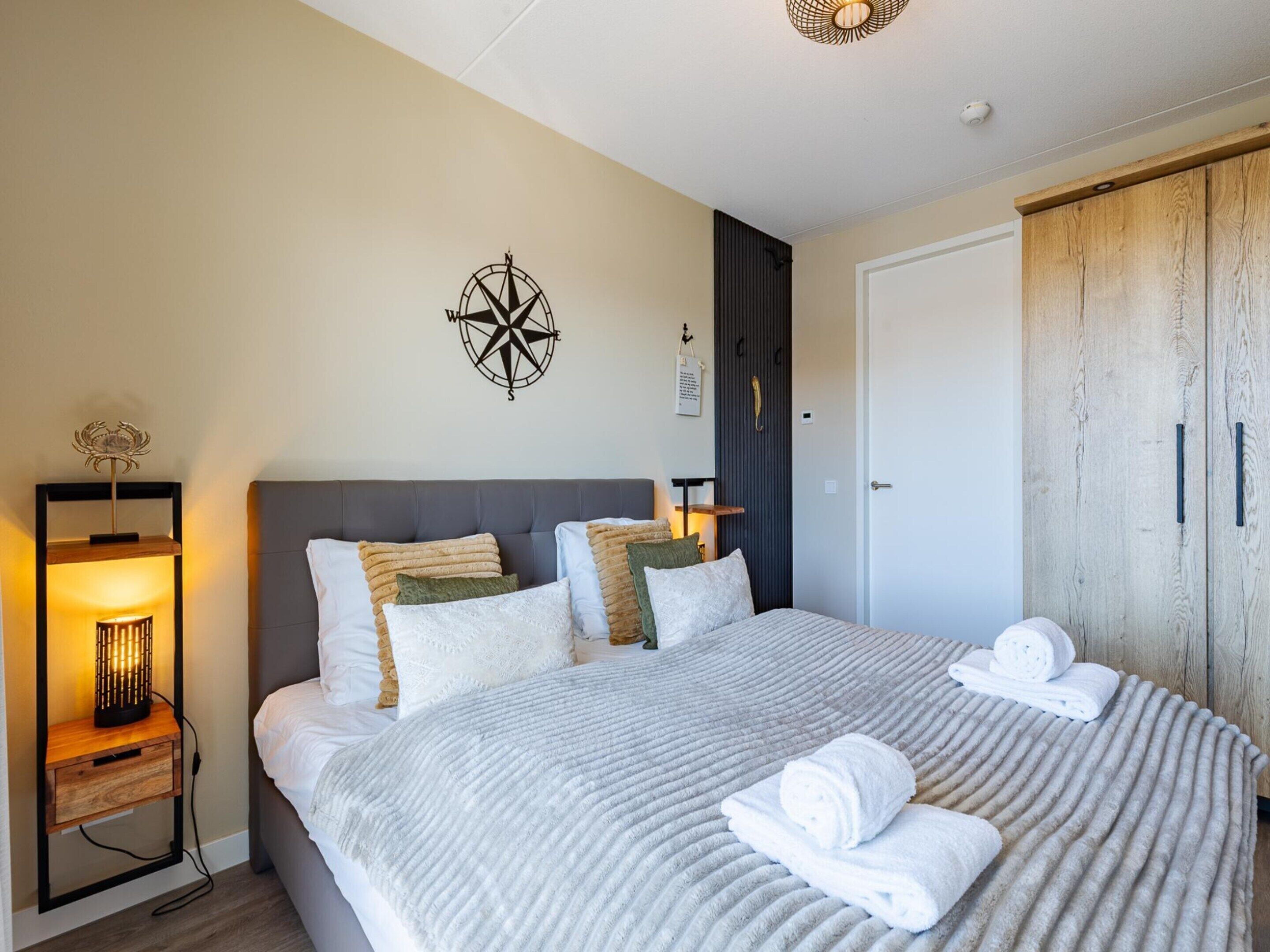 A neatly arranged bedroom with a large bed, a bedside table with a lamp, a wardrobe, and a wall-mounted compass decoration.