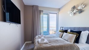 A hotel room with a bed, a TV mounted on the wall, a window with curtains, and a view of the sea. - Apartment in Sint-annaland With Beach Access (Sint-Annaland)