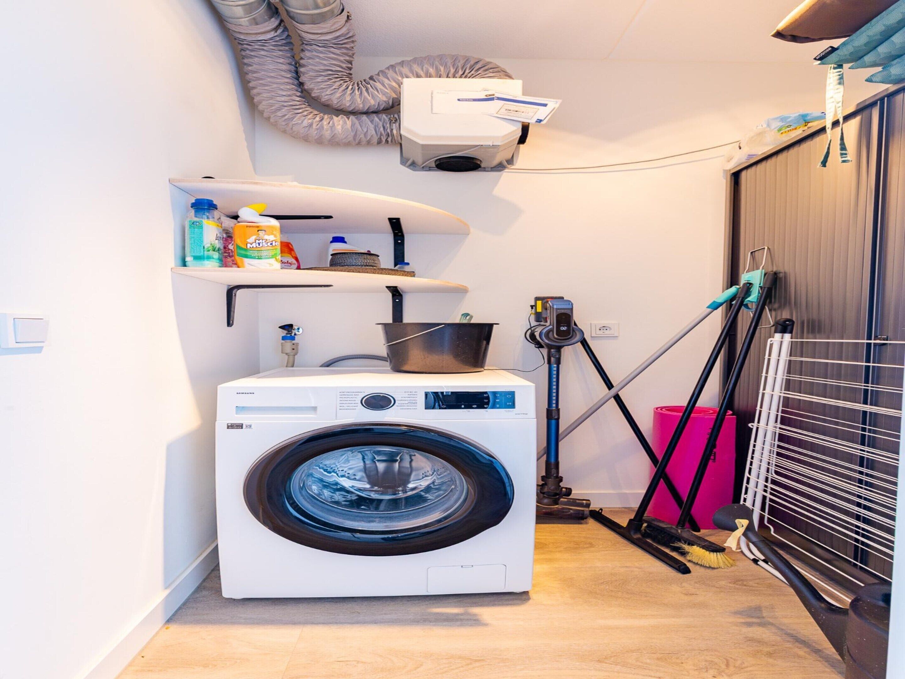 A compact laundry area with a front-loading washing machine, a shelf with cleaning supplies, and a drying rack.