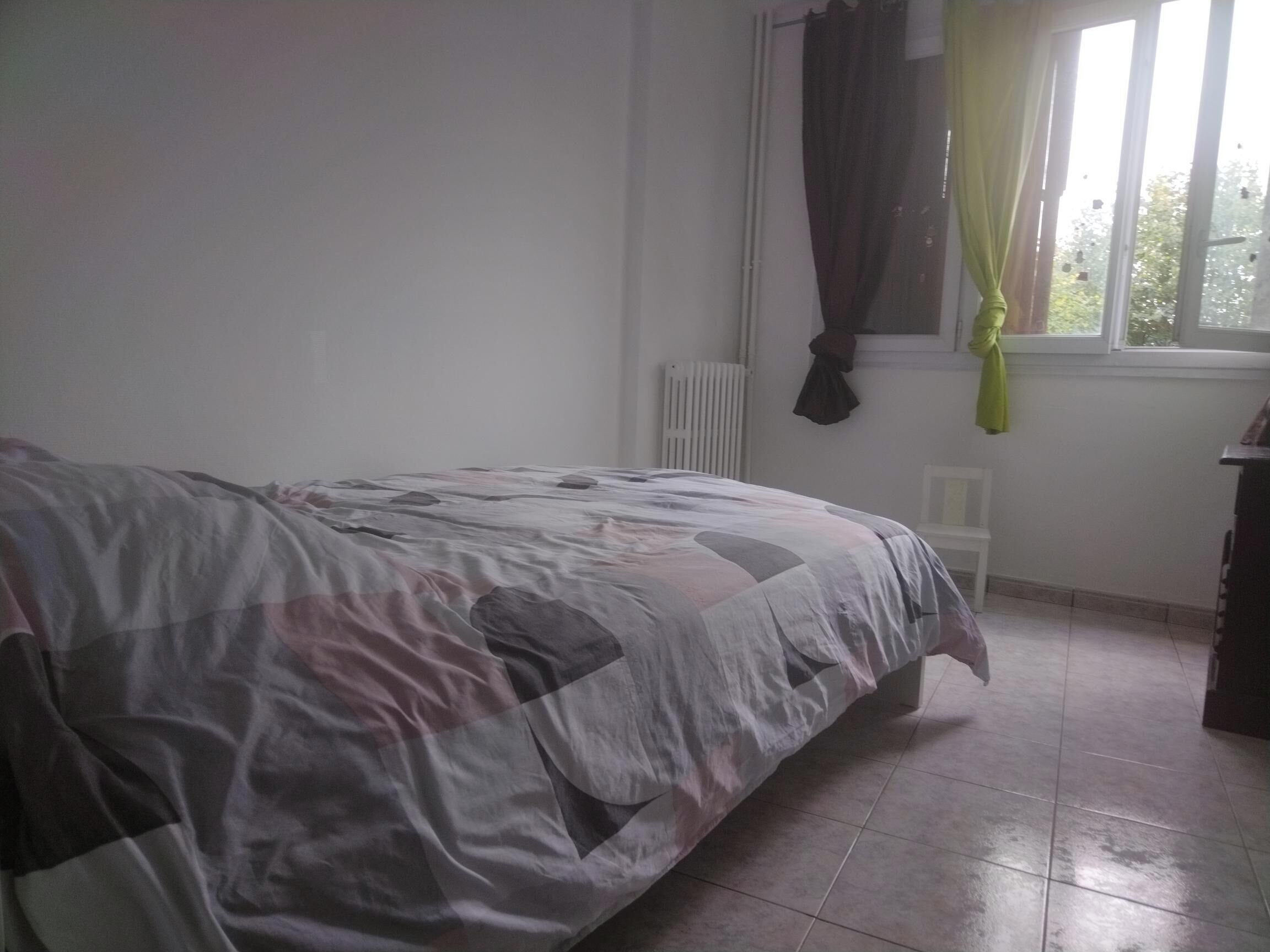 A bedroom with a bed, a radiator, a window with curtains, and a chair.