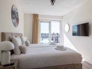 Habitación de hotel moderna con una cama grande, un televisor de pantalla plana, un cuadro y vistas a la ciudad desde la ventana.