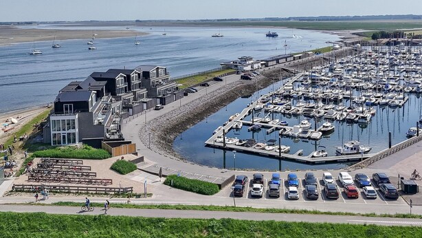 Marina con numerosas embarcaciones amarradas, zona de estacionamiento con coches y un edificio de diseño moderno cerca del agua.
