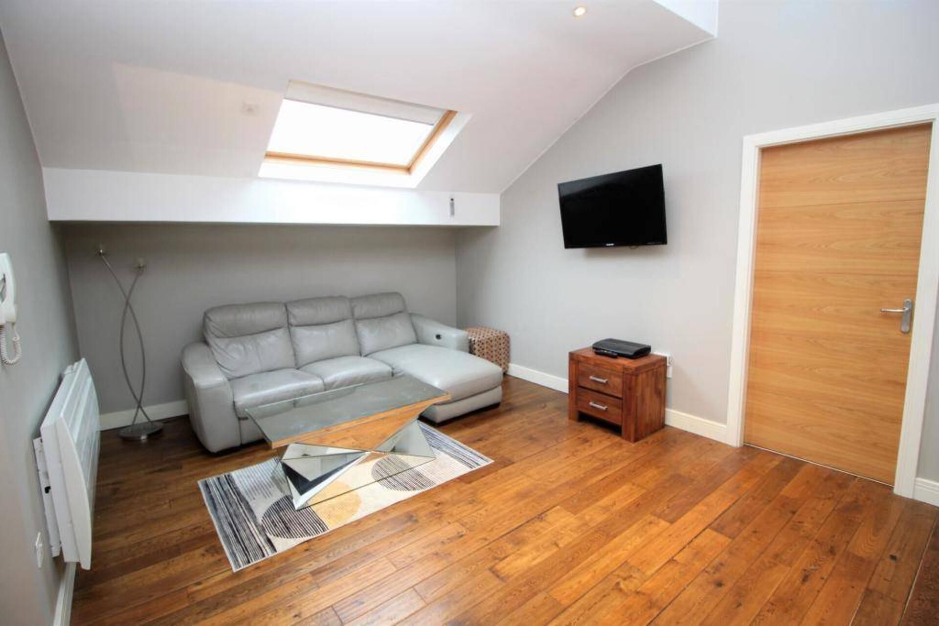 A room with a sloped ceiling, a grey sofa, a wooden floor, a TV mounted on the wall, and a door.