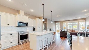 A modern kitchen with white cabinetry, a central island with bar stools, and a dining area with a table and chairs. - Mimi`s Cottage - Golf Cart Included (Osoyoos)