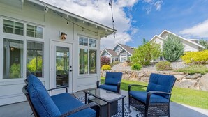 A patio with wicker furniture and a glass table, a white house with large windows, and a landscaped garden. - Mimi`s Cottage - Golf Cart Included (Osoyoos)