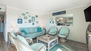Una sala de estar con un sofá color turquesa, sillas de mimbre, una mesa de centro y un letrero que dice 'Seas the Bay'.
