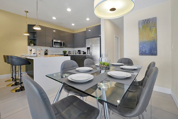 Una cocina moderna con gabinetes grises, electrodomésticos de acero inoxidable y un comedor con mesa de vidrio y sillas grises.