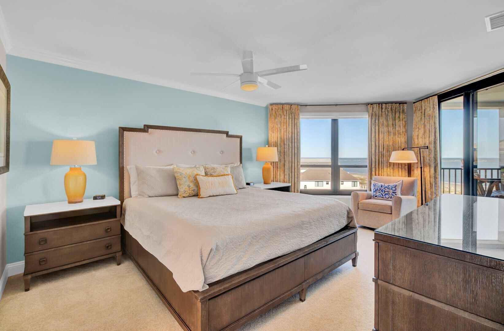 A bedroom with a large bed, two bedside tables with lamps, a nightstand, a dresser, a chair, and a balcony with a view.