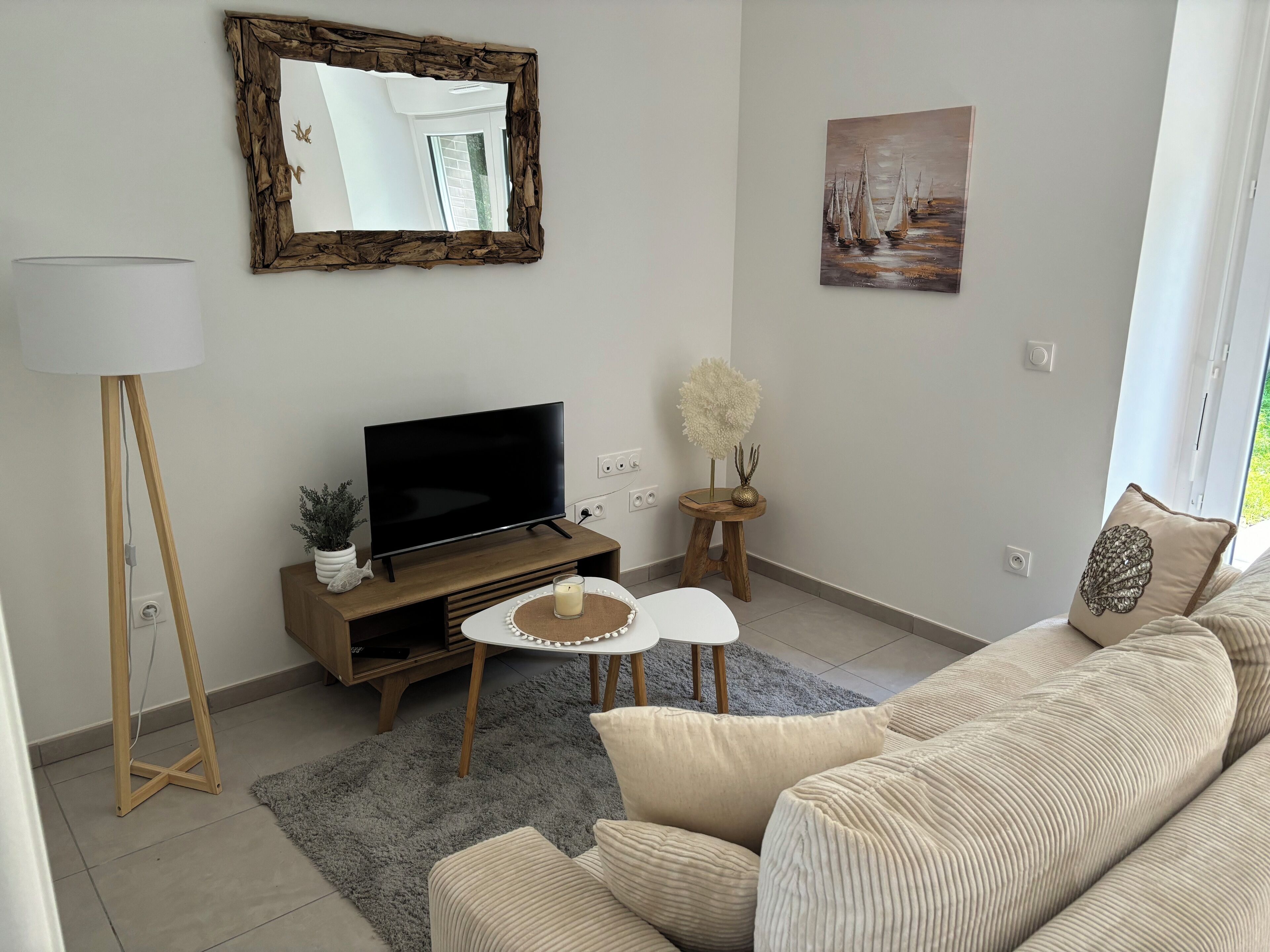 Uma sala de estar com televisão de tela plana sobre um suporte de madeira, um sofá, um abajur, uma pequena mesa com uma vela e um quadro na parede.