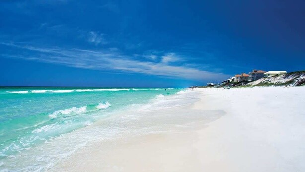 Une plage de sable blanc bordée d’une eau turquoise. On aperçoit des maisons sur le côté droit de l’image.
