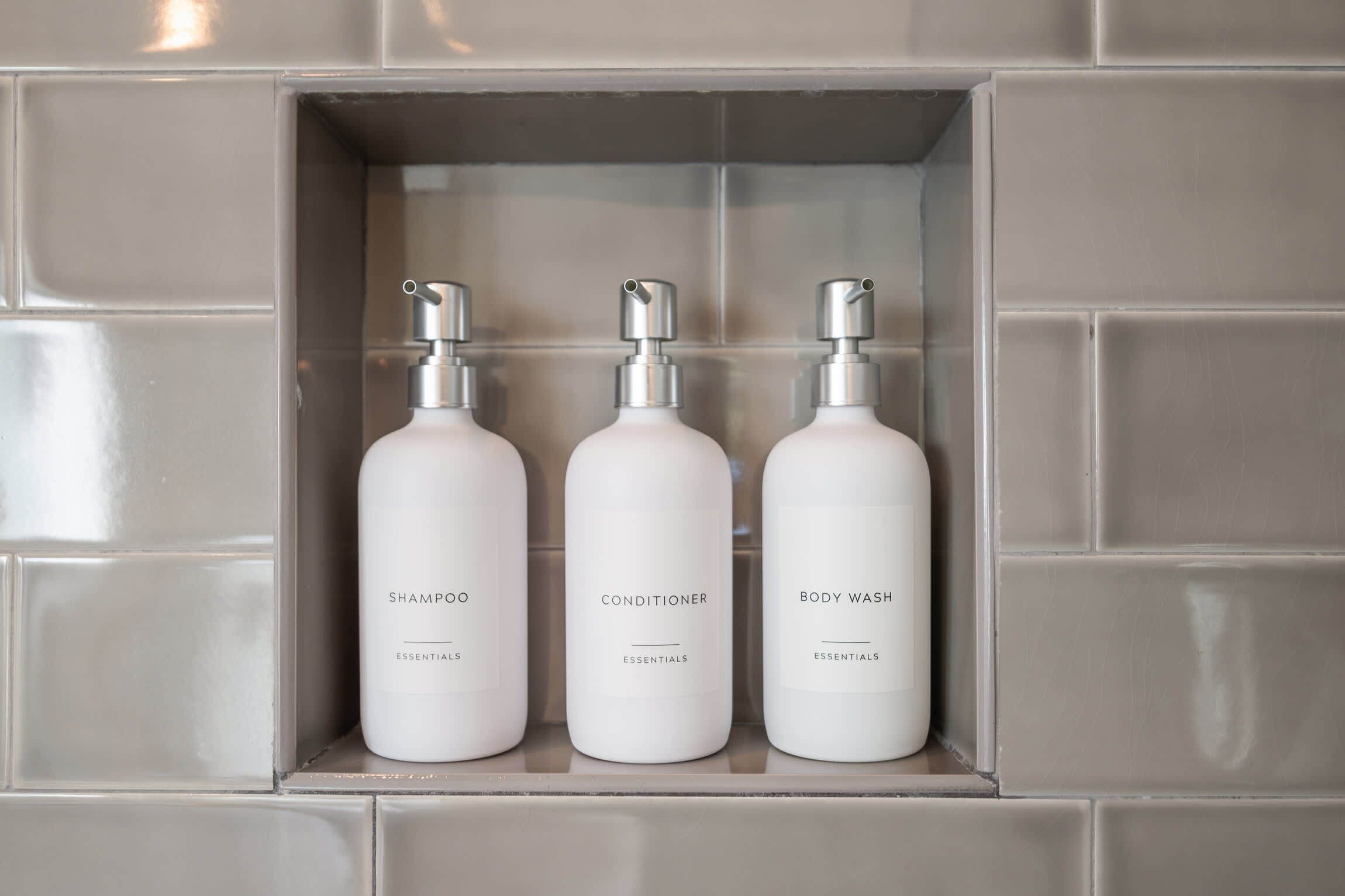 Tres botellas blancas etiquetadas como champú, acondicionador y gel de baño en un armario de azulejos.