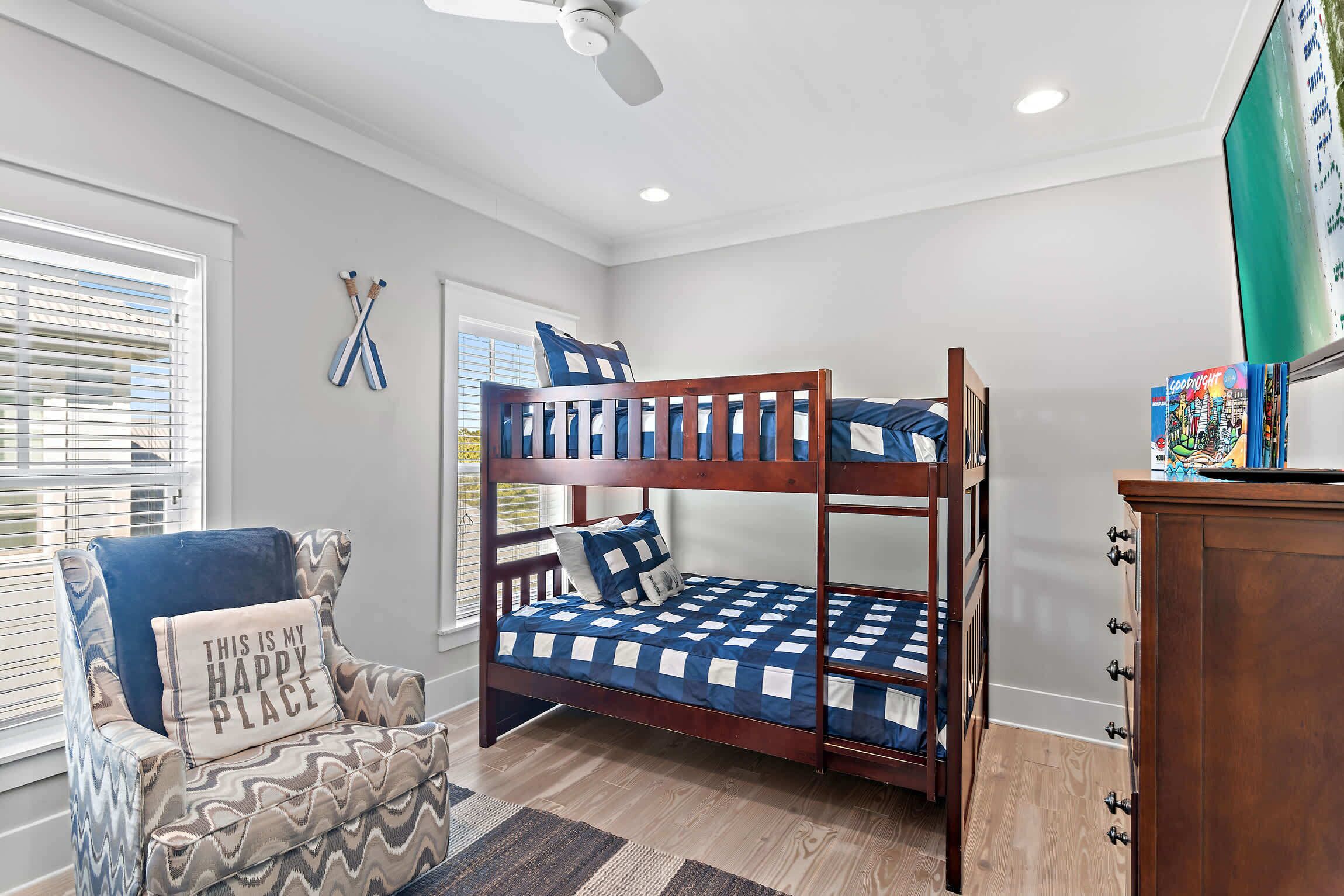 A bunk bed with a checkered blue and white bedding, a chair with a 'This is my happy place' pillow, and a ceiling fan.