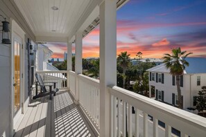 A wooden porch with a railing, a bench, and a view of a cityscape and palm trees during sunset.