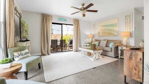 A spacious living room with a sofa, a coffee table, a TV, and a ceiling fan. - 3BR Cocoa Beach Oasis | 1 Block to Beach (Cocoa Beach)
