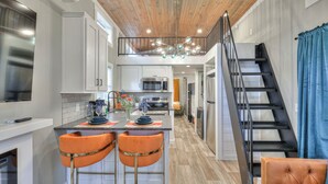 A modern kitchen with white cabinets, a central island with orange chairs, a stainless steel refrigerator, and a wooden ceiling.