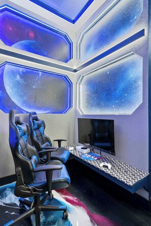 A gaming setup with two chairs, a desk, a monitor, and a gaming console.