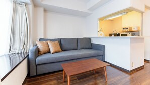 Ein modernes Wohnzimmer mit grauem Sofa, zwei Lederkissen, einem Holztisch und einer Küchenzeile mit Mikrowelle und Toaster.