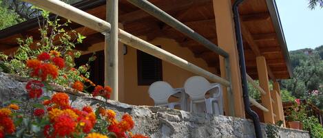 A rustic building with a stone wall and a wooden roof. There are white plastic chairs and a stone wall with flowers in the foreground.