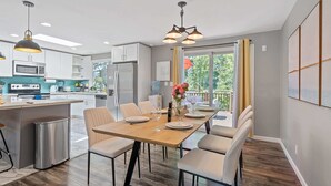 A modern kitchen with white cabinets, a central island, and a dining area with a wooden table and chairs. - Chic Kirkland Home Overlooking Windsor Vista Park (Kirkland)