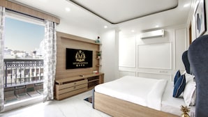 A modern hotel room with a large bed, a TV mounted on a wooden panel, a balcony with a view of buildings, and an air conditioning unit on the wall. - MIYABI HOTEL (Gurugram)