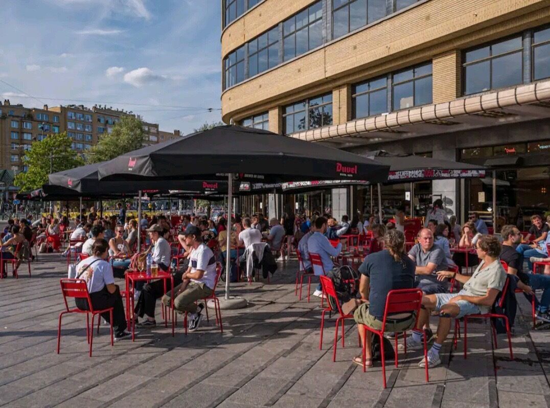 Un espace extérieur aménagé avec des chaises et des tables rouges, un parasol noir, et un bâtiment avec de nombreuses fenêtres en arrière-plan.