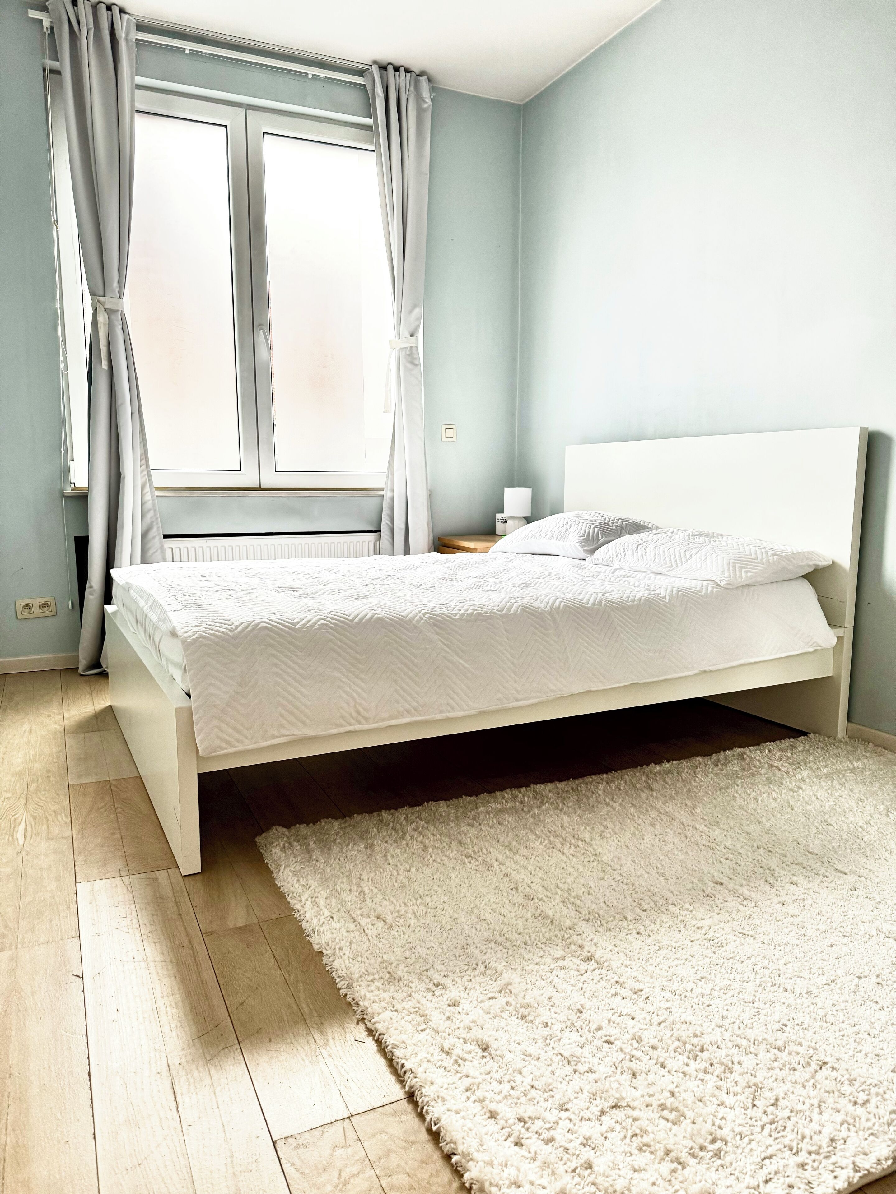 Une chambre à coucher avec un lit, une table de chevet, une fenêtre avec des rideaux et un tapis blanc.